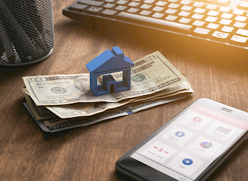 Blue plastic house figure on top of a stack of money next to a phone that has an online banking app open